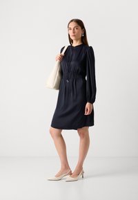 Navy blue dress with a zipper and gathered waist, long sleeves, paired with a beige tote bag and neutral slingback heels.