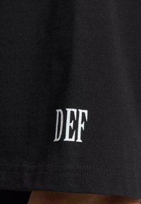 Tricou negru cu un finisaj texturat, având literele albe imprimate "DEF" aproape de tiv, cu o croială dreaptă și detalii de cusătură vizibile.