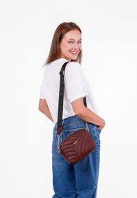 Hnědá prošívaná crossbody taška s černým logem na popruhu, se zlatým akcentem a obdélníkovým tvarem. Nosená přes rameno.