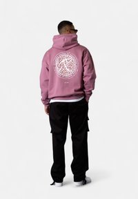 Pink hoodie with a round white leopard-print graphic on the back, paired with black pants and white sneakers. Relaxed fit and soft fabric.