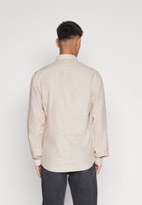 Camicia beige a maniche lunghe con colletto button-down, tessuto testurizzato e polsini con un bottone, vista da dietro su uno sfondo semplice.
