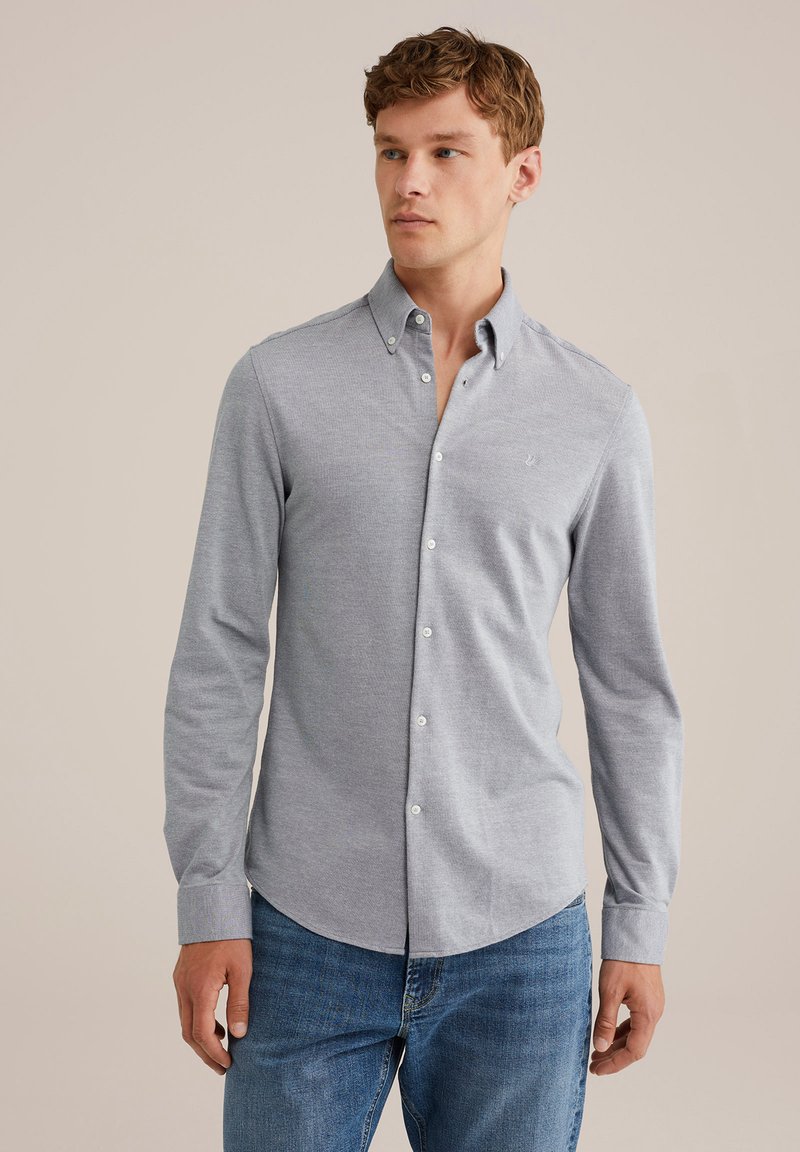 Hombre vestido con una camisa de botón gris claro con cuello abotonado y jeans azules, de pie contra un fondo beige liso.