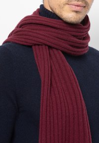 Burgundivärinen neulehuivi, jossa on uritettu pinta, laskeutuu kaulan ympäri ja yhdistyy tummansiniseen neulepuseroon. Pehmeä ja lämmin ilme.