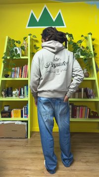 Beige hoodie with "Pegador" text and logo on the back, paired with loose blue jeans. Bright yellow wall and green shelves in background.