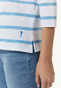 Weiß gestreiftes Langarmshirt mit blauen horizontalen Streifen, seitlichen Schlitzen und gesticktem Logo. Getragen mit hellblauen Jeans.