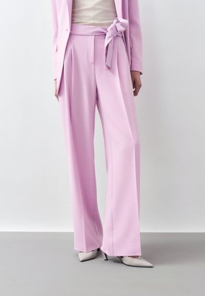 Kobieta ubrana w pastelowe różowe spodnie o szerokich nogawkach z paskiem wiązanym w talii, biały top, pasująca pastelowo różowa marynarka oraz białe szpiczaste szpilki.