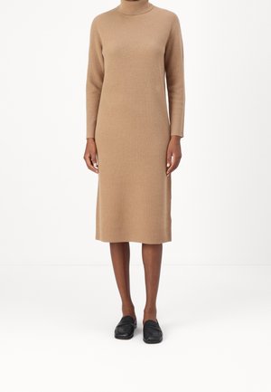 Persona che indossa un lungo vestito maglione a coste colore cammello abbinato a mocassini neri senza lacci, in piedi davanti a uno sfondo bianco neutro.