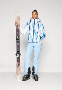 Skijacke in Weiß mit blauen Wellenmustern, kombiniert mit hellblauen Skihosen. Ein Ski wird in einer Hand gehalten, mit schwarzen Bindungen und einer strukturierten Oberfläche.