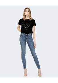 Musthave must kas t-särk kolmnurkse graafilise logoga, millele on sobitatud helesinised kitsad teksad ja kehavärvi kõrged kontsad. Ühtlane taust.
