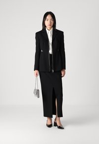 Crni krojeni blazer s patent zatvaranjem, u kombinaciji s crnom suknjom visokog proreza. Dodaci uključuju malu torbicu s kariranim uzorkom i sjajne crne štikle.