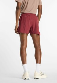 Pantaloncini sportivi bordeaux con tessuto leggero e liscio, vita elastica e spacchi laterali per maggiore flessibilità, abbinati a scarpe da corsa color crema.
