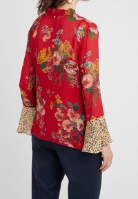 Blusa floral roja con mangas largas, que presenta una tela transparente y puños con estampado de leopardo. Incluye detalles de botones en la parte trasera.