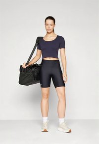 Sorte treningsshorts og en marineblå crop-top, kombinert med en stor svart duffelbag. Lyse sneakers med hvite sokker. Enkel design.