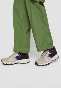 Lekkie buty outdoorowe z beżową cholewką, fioletowymi akcentami, czarnym wzmocnieniem pięty i nakrapianą podeszwą; zaprojektowane z myślą o stabilności i przyczepności.