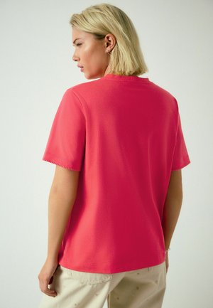 Trumpomis rankovėmis, apvalia iškirpte marškinėliai iš ryškiai rožinės medžiagos. Turi laisvą kirpimą ir matomas siuvimo detales ant rankovių.