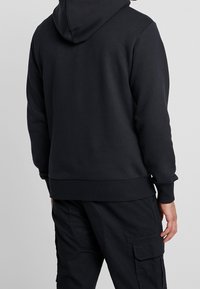 Uomo con un hoodie nero e pantaloni cargo neri di spalle, in piedi su uno sfondo grigio chiaro uniforme.