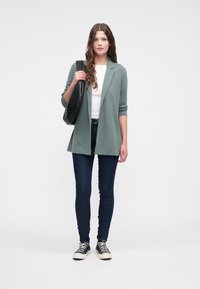 Mintgrønn blazer over en hvit topp, mørke skinny jeans og svarte sneakers. Svart skulderveske, bølgete hår, nøytral bakgrunn.