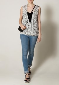 Blusa senza maniche bianca e nera con motivo floreale, abbinata a jeans blu aderenti e sandali neri. La blusa presenta un design a scollo a V profondo.