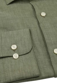 Olijfgroene linnen blouse met een puntige kraag, twee knoopmanchetten en witte knopen. De stof heeft een gestructureerde afwerking en een subtiele weving.