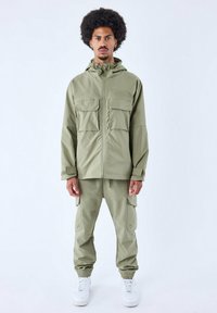 Set di giacca e pantaloni di un verde oliva chiaro realizzato in tessuto strutturato, caratterizzato da tasche frontali, cappuccio e polsini elastici, abbinato a scarpe bianche.