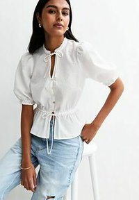 Kvinna med långt mörkt hår som bär en vit blus med puffärmar och snörning samt ljusblå slitna jeans, sittande på en vit pall.