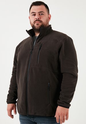 Chaqueta de forro polar marrón oscuro con un cuello alto, un bolsillo en el pecho con cremallera y dos bolsillos laterales con cremallera. Textura suave y diseño ajustado.