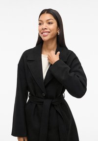 Smilende kvinne med langt mørkt hår har på seg en svart beltesteejakke over en hvit topp, og holder kragen på jakken med den ene hånden.