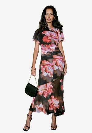 Robe maxi fleurie en tissu noir transparent avec de grandes fleurs roses. Présente des manches courtes en volant et une silhouette ajustée. Accompagnée d'un sac à main vert.