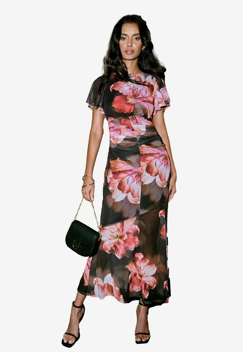Robe maxi fleurie en tissu noir transparent avec de grandes fleurs roses. Présente des manches courtes en volant et une silhouette ajustée. Accompagnée d'un sac à main vert.