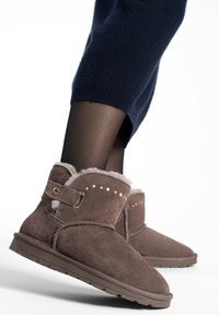 Botas de tobillo de ante marrón con forro de pelo, que presentan tachuelas doradas y una correa lateral. Suelas planas y punteras redondeadas.