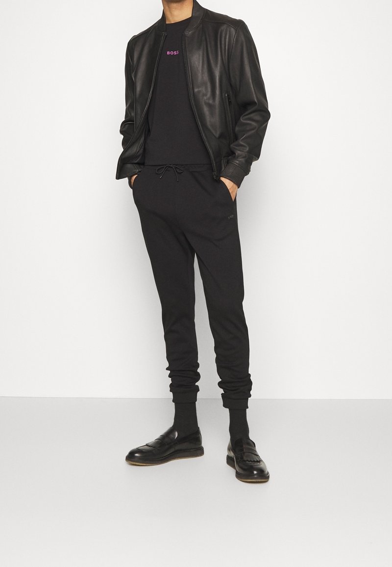 Svart läderbomberjacka över en svart T-shirt med "BOSS"-logotyp, tillsammans med avslappnade svarta joggers och svarta loafers.