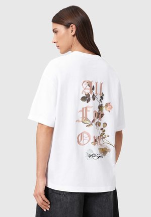 Woman with straight brown hair wearing a white oversized T-shirt with "All For One" text and floral design on the back, paired with black jeans.