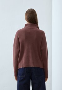 Brauner Strickpullover mit hohem Kragen, geripptem Muster und überschnittenen Schultern. Kombiniert mit dunkelblauen Jeans.