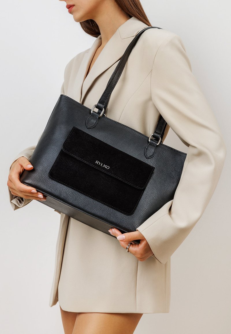 Woman in beige blazer holding large black leather tote bag with front suede pocket and "RYŁKO" logo.