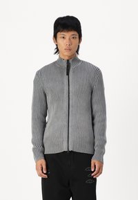Suéter gris de canalé con cremallera y cuello alto, con una cremallera negra en contraste. Presenta rayas verticales y mangas ajustadas, diseño entallado.