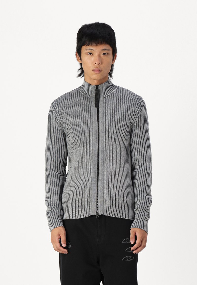 Suéter gris de canalé con cremallera y cuello alto, con una cremallera negra en contraste. Presenta rayas verticales y mangas ajustadas, diseño entallado.