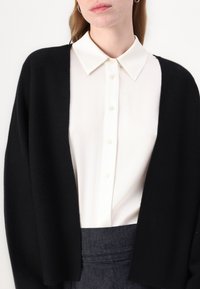 Černý cardigan s otevřeným předním dílem, měkká textura, nošený přes bílou košili s knoflíky a špičatým límečkem. Viditelné tmavě šed�é kalhoty na míru.