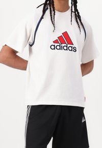 Bílá bavlněná tričko s krátkými rukávy, se červeným a tmavě modrým logem "adidas" a akcenty podél ramen. Doplněno černými šortkami.