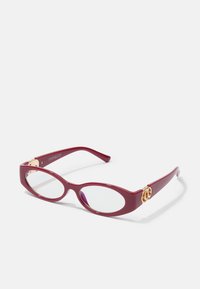 Červené plastové brýle ve tvaru oválu s průhlednými čočkami. Obsahují zlaté kovové detaily na spáncích a mají elegantní moderní design.
