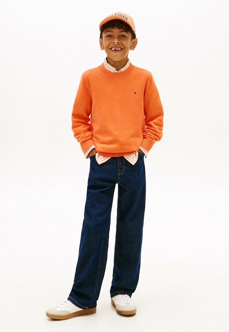 Ragazzo che sorride con le mani nelle tasche, indossa un maglione arancione, un berretto abbinato, jeans blu scuro, sneakers bianche e una camicia a righe.