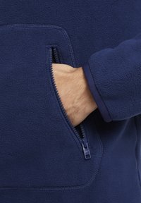 Giacca in pile blu navy con tasca con zip e texture morbida. Caratterizzata da un colletto alto e dettagli a contrasto sui polsini.