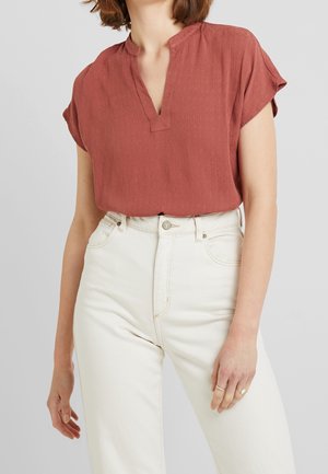 Femme portant un chemisier rouge rouille à manches courtes rentré dans un jean taille haute blanc cassé sur fond uni.