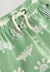 Groene zwembroek met witte dierenprints, waaronder zebra's en krokodillen. Elastische tailleband met witte trekkoorden en zwarte oogdetail.