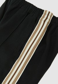 Pantalon de survêtement noir avec une large ceinture élastique et une bande latérale ornée de trois lignes horizontales en beige et marron. Texture lisse.
