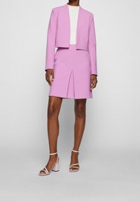 Růžový na míru šitý blazer a mini sukně, obsahující zkrácenou bundu bez zapínání a sukni s předním rozparkem. Doplněno o světle růžové lodičky.