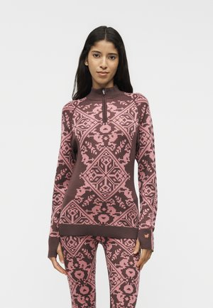 Femme portant un pull zippé marron et rose à motifs et des leggings assortis, debout devant un fond clair uni.