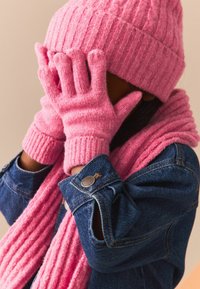Roze gebreide beanie en handschoenen met ribbeltextuur, gecombineerd met een bijpassende sjaal. Blauwe spijkerjack met een knoopdetail op de manchetten.