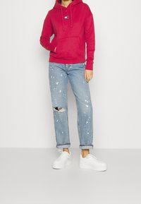 Červená mikina s kapucí, přední kapsou a šňůrkou, v kombinaci se světle modrými roztrhanými džíny s flekatým designem. Spárované s bílými teniskami.