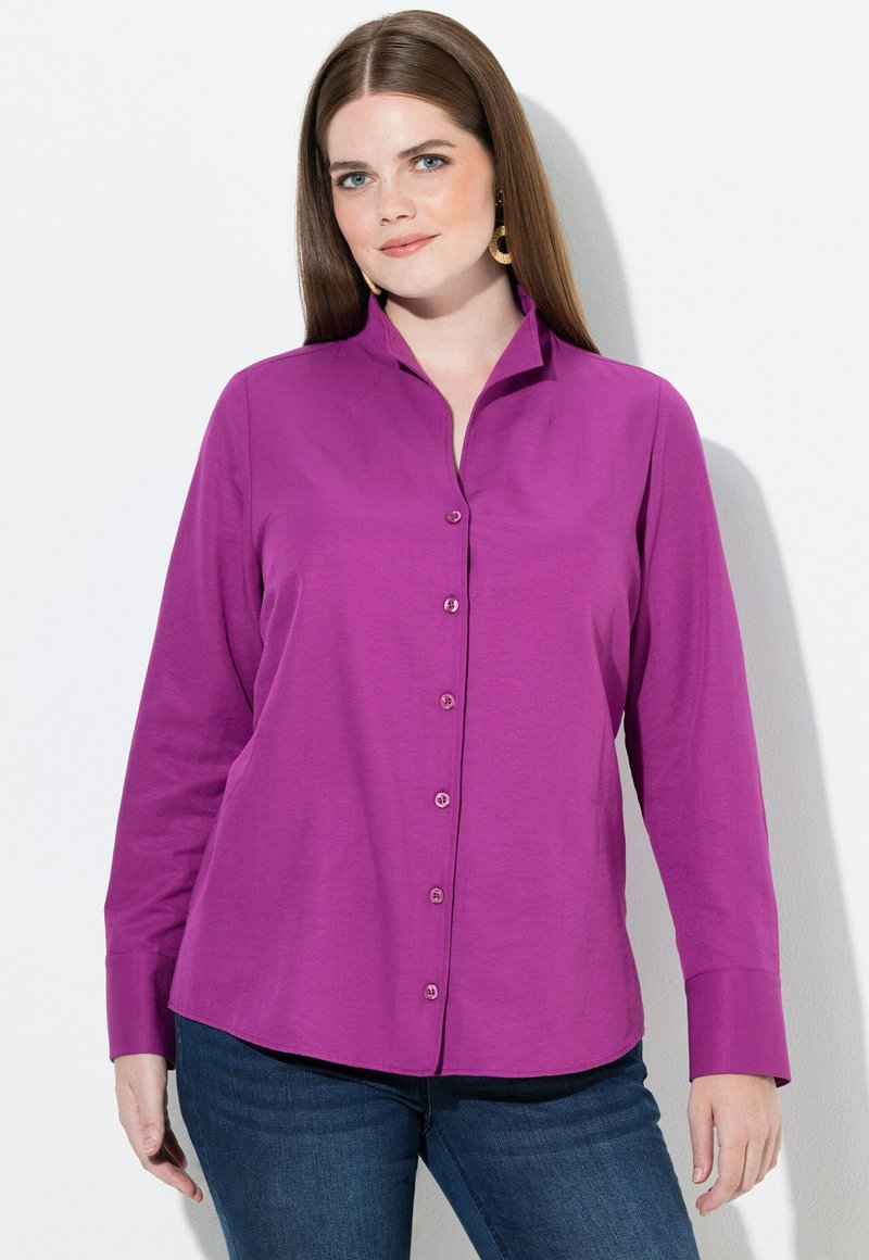 Camicia button-up viola con colletto, maniche lunghe e texture morbida. Presenta bottoni abbinati e un'orlo leggermente curvato. Indossata con jeans.