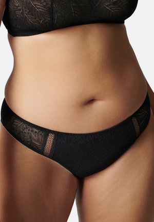 Kobieta w czarnym koronkowym biustonoszu i dopasowanej bieliźnie, tors i biodra widoczne na jednolitym tle.